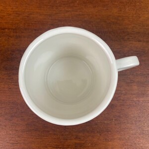 K&ouml;nnte beinhalten: Eine wei&szlig;e Keramik-Tasse mit Henkel, von oben gesehen. Das Innere der Tasse ist hellcremefarben. Die Tasse steht auf einer dunkelbraunen Holzoberfl&auml;che.
