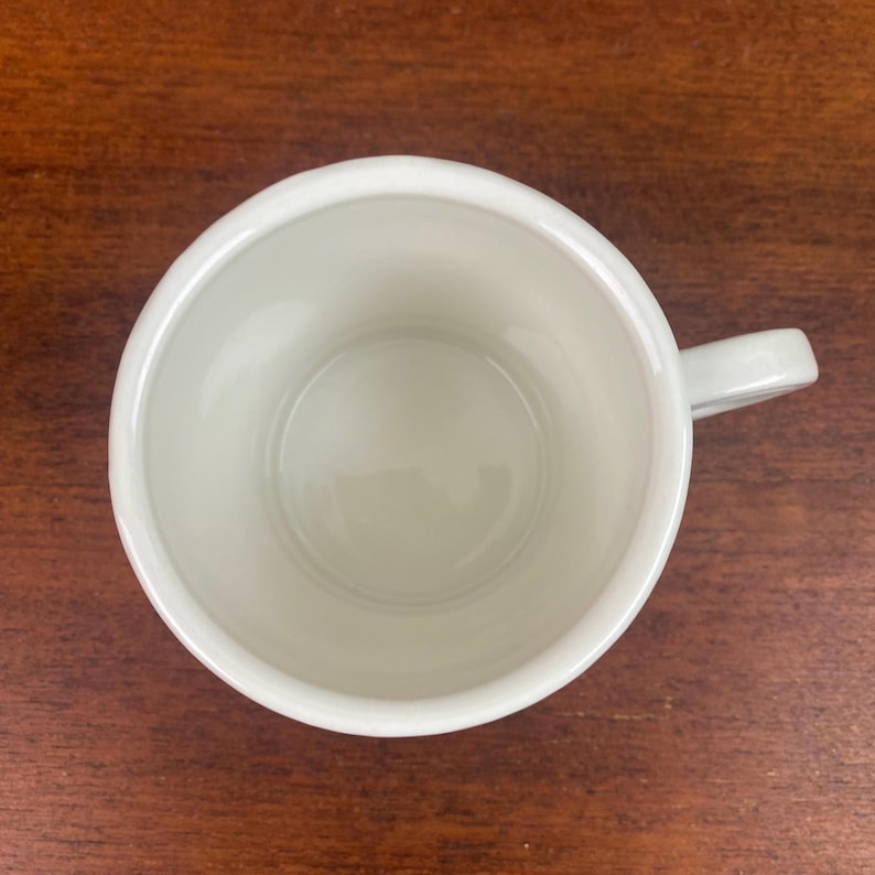 K&ouml;nnte beinhalten: Eine wei&szlig;e Keramik-Tasse mit Henkel, von oben gesehen. Das Innere der Tasse ist hellcremefarben. Die Tasse steht auf einer dunkelbraunen Holzoberfl&auml;che.