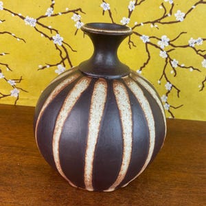 May include: A dark brown ceramic vase with vertical cream-colored stripes. The vase has a rounded body and a narrow neck. The background features a yellow surface with a floral pattern.