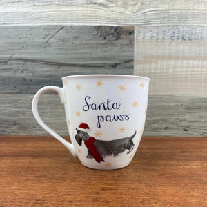 May include: White ceramic mug with a black and gray dog wearing a Santa hat and red scarf. The mug has the words "Santa paws" in blue script, with yellow snowflakes. The mug has a curved handle.