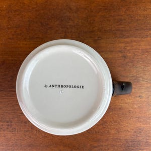 May include: White ceramic mug with a black handle. The bottom of the mug has the text "by ANTHROPOLOGIE" printed in black.