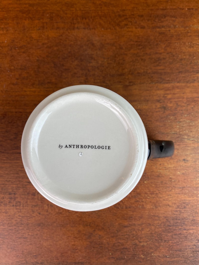 May include: White ceramic mug with a black handle. The bottom of the mug has the text "by ANTHROPOLOGIE" printed in black.