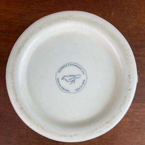 K&ouml;nnte beinhalten: Boden einer cremewei&szlig;en Keramik-Tasse mit einem kreisf&ouml;rmigen blauen Stempel, der eine Vogelsilhouette und den Text "MICROWAVE & DISHWASHER SAFE" und "MADE IN CHINA" zeigt. Die Tasse hat eine leicht strukturierte Oberfl&auml;che.