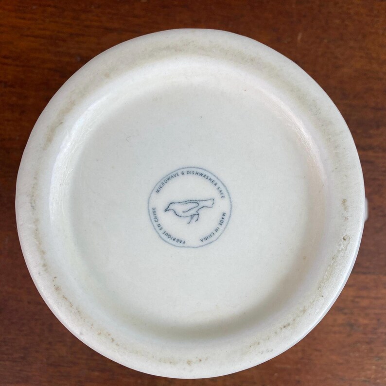 K&ouml;nnte beinhalten: Boden einer cremewei&szlig;en Keramik-Tasse mit einem kreisf&ouml;rmigen blauen Stempel, der eine Vogelsilhouette und den Text "MICROWAVE & DISHWASHER SAFE" und "MADE IN CHINA" zeigt. Die Tasse hat eine leicht strukturierte Oberfl&auml;che.