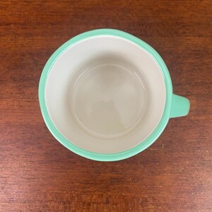 May include: Overhead view of a ceramic mug with a mint green rim and handle. The interior of the mug is off-white. The mug sits on a dark brown wooden surface.