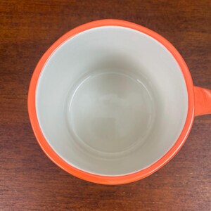 May include: Overhead view of a white ceramic mug with an orange rim and handle. The mug is empty and sits on a dark wooden surface. The interior of the mug is white, and the rim and handle are a bright orange.