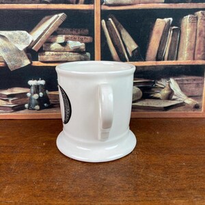 May include: A white ceramic mug with a rounded base and a handle. The mug has a black circular design on the side. The background shows a wooden shelf filled with books.