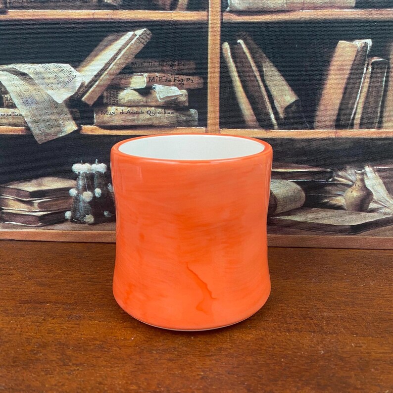 May include: A cylindrical orange ceramic mug with a white interior. The mug has a glossy finish and a slightly irregular shape. The background features a wooden surface and a bookshelf with books.