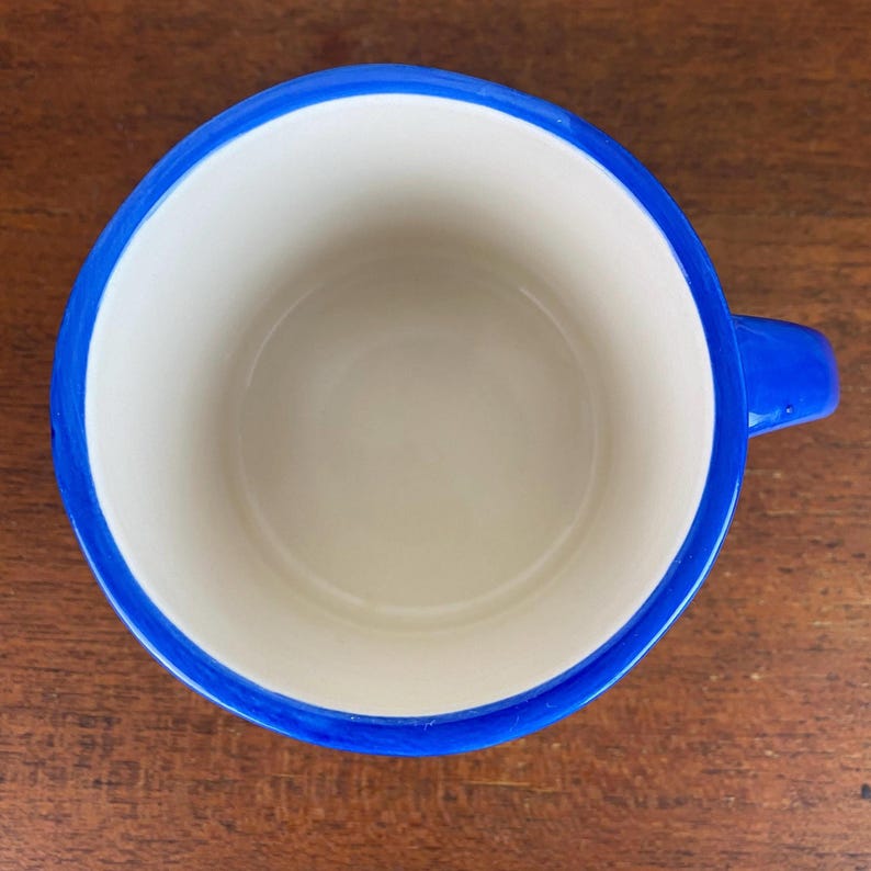 May include: A ceramic mug viewed from above, featuring a bright blue rim and handle. The interior is white, creating a contrast with the blue. The mug is set on a wooden surface.