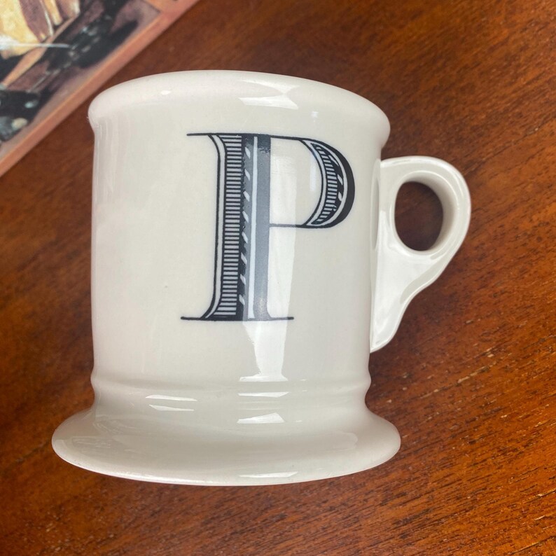 May include: A white ceramic mug featuring a large black letter "P". The letter is decorated with a detailed design. The mug has a handle and a wide base. The mug is on a wooden surface.