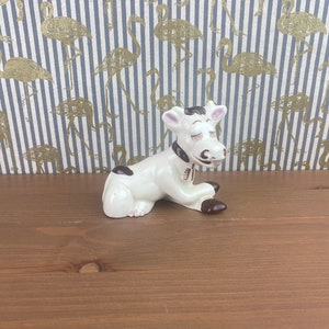 Op de afbeelding: Een wit keramiek koeienbeeldje met bruine vlekken en gesloten ogen. De koe zit met zijn hoofd naar beneden gekanteld en zijn poten onder zijn lichaam gevouwen.