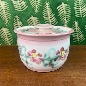 以下が含まれることがあります： ピンク色のセラミックボウルと蓋。手描きのピンクの花と緑のつるで飾られています。ボウルは薄いピンク色の縁と白いベースです。背景には、白いシダの葉の緑色のパターンがあります。