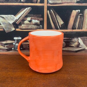 May include: An orange ceramic mug with a white interior. The mug has a curved handle and a smooth, glossy finish. The background features a bookshelf with books.