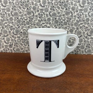 K&ouml;nnte beinhalten: Wei&szlig;e Keramiktasse mit einem schwarzen Buchstaben "T"-Design. Die Tasse hat einen abgerundeten Boden und einen Henkel. Der Hintergrund zeigt ein Blumenmuster.