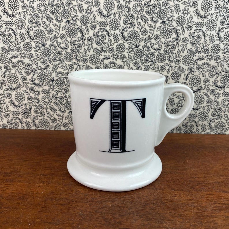 K&ouml;nnte beinhalten: Wei&szlig;e Keramiktasse mit einem schwarzen Buchstaben "T"-Design. Die Tasse hat einen abgerundeten Boden und einen Henkel. Der Hintergrund zeigt ein Blumenmuster.