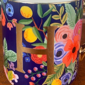 May include: A ceramic mug with a navy blue background, adorned with a gold letter "H" and a vibrant floral pattern. The design includes orange citrus fruits, and colorful flowers in shades of red, pink, and blue. The mug has a gold rim and handle.