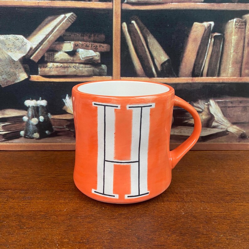 May include: Orange ceramic mug with a white interior and a large white letter "H" in the center. The mug has a curved handle and is set against a blurred background of books.