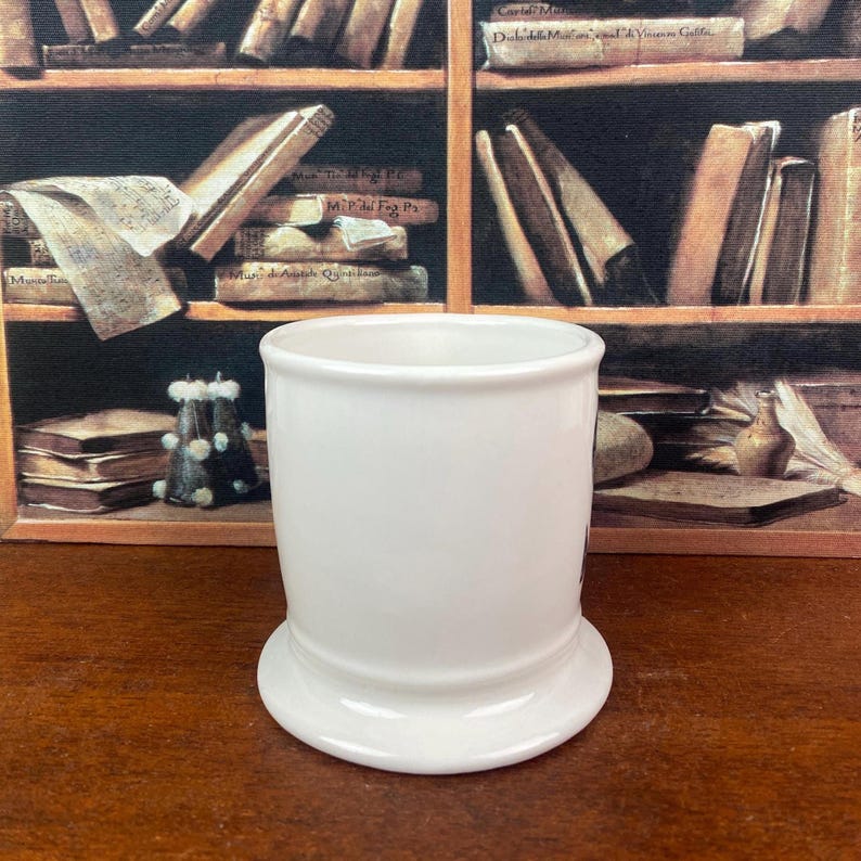 May include: A white ceramic mug with a rounded base and a cylindrical body. The mug has a glossy finish and is set against a backdrop of a bookshelf filled with books. The mug is empty and ready to be filled.