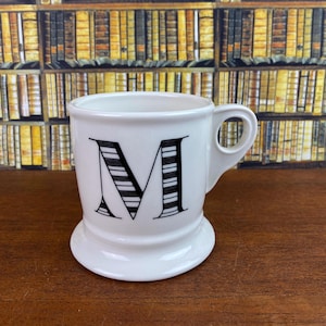 May include: White ceramic mug with a large black and white striped letter "M" on the front. The mug has a rounded base and a small handle. The background features a wall of books.
