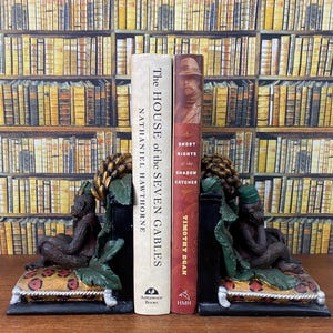 May include: A pair of bookends featuring a monkey sitting on a cushion. The bookends are holding up two books, "The House of the Seven Gables" by Nathaniel Hawthorne and "Short Nights of the Shadow Catcher" by Timothy Egan.