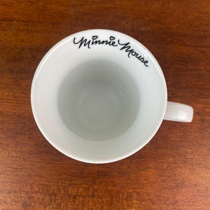 Puede incluir: Taza de cer&aacute;mica blanca con asa, con la inscripci&oacute;n "Minnie Mouse" en negro. La taza se encuentra sobre una superficie de madera marr&oacute;n, mostrando su dise&ntilde;o limpio y sencillo. El interior de la taza es visible.