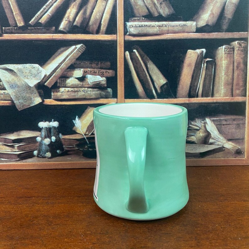 May include: A light green ceramic mug with a white interior and a curved handle. The mug is set against a backdrop of a wooden bookshelf filled with books. The mug is on a wooden surface.