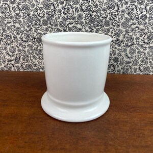 K&ouml;nnte beinhalten: Ein wei&szlig;er Keramikbecher mit rundem Boden und zylindrischem K&ouml;rper. Der Becher steht vor einem floral gemusterten Hintergrund und ruht auf einer Holzoberfl&auml;che. Der Becher ist etwa 10 cm hoch.