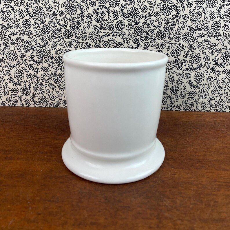 K&ouml;nnte beinhalten: Ein wei&szlig;er Keramikbecher mit rundem Boden und zylindrischem K&ouml;rper. Der Becher steht vor einem floral gemusterten Hintergrund und ruht auf einer Holzoberfl&auml;che. Der Becher ist etwa 10 cm hoch.