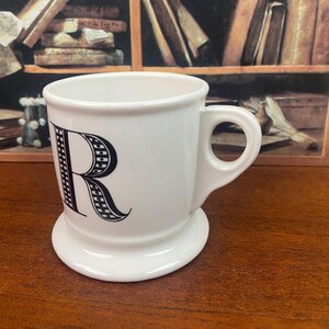 K&ouml;nnte beinhalten: Wei&szlig;e Keramiktasse mit einem gro&szlig;en schwarzen Buchstaben "R" auf der Vorderseite. Die Tasse hat einen abgerundeten Boden und einen Henkel. Der Hintergrund umfasst eine Holzoberfl&auml;che und ein B&uuml;cherregal.