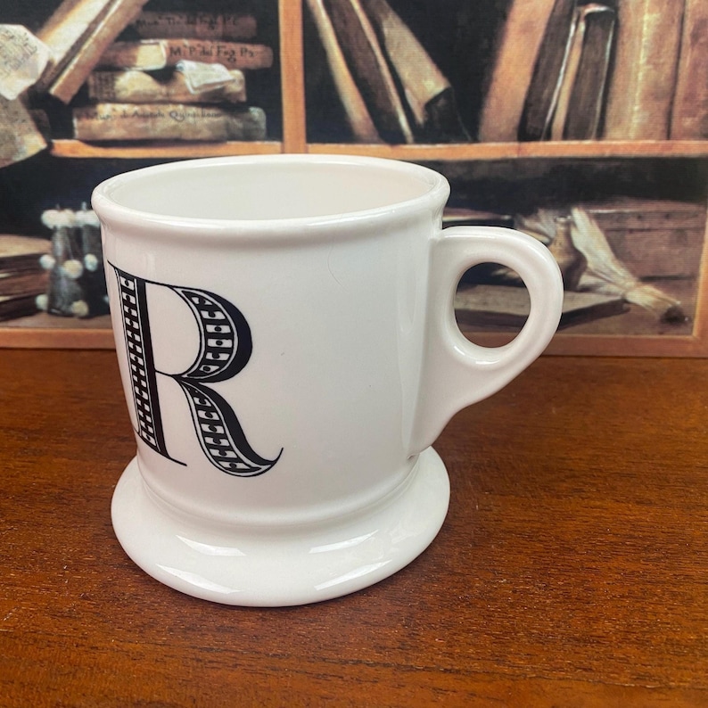K&ouml;nnte beinhalten: Wei&szlig;e Keramiktasse mit einem gro&szlig;en schwarzen Buchstaben "R" auf der Vorderseite. Die Tasse hat einen abgerundeten Boden und einen Henkel. Der Hintergrund umfasst eine Holzoberfl&auml;che und ein B&uuml;cherregal.