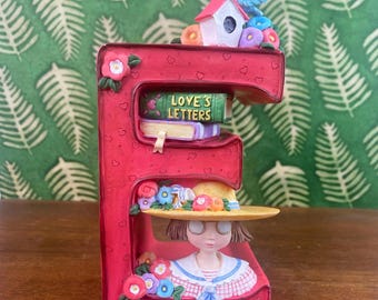 Puede incluir: Figurita decorativa roja con la letra "E" con una niña con sombrero, libros con "Love's Letters", una casita para pájaros y un pájaro azul. La figurita está decorada con flores y corazones.