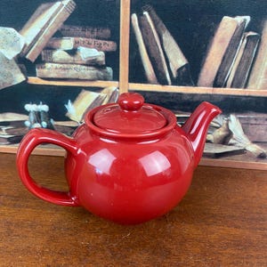 Può includere: Una teiera in ceramica rossa lucida con coperchio abbinato e manico curvo. La teiera ha un corpo arrotondato e un beccuccio per versare. Lo sfondo presenta una mensola con libri.