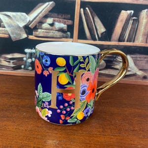 May include: A ceramic mug with a gold handle and the letter "H" in gold. The mug is decorated with a floral pattern in shades of blue, orange, pink, and green. The mug is sitting on a wooden surface.