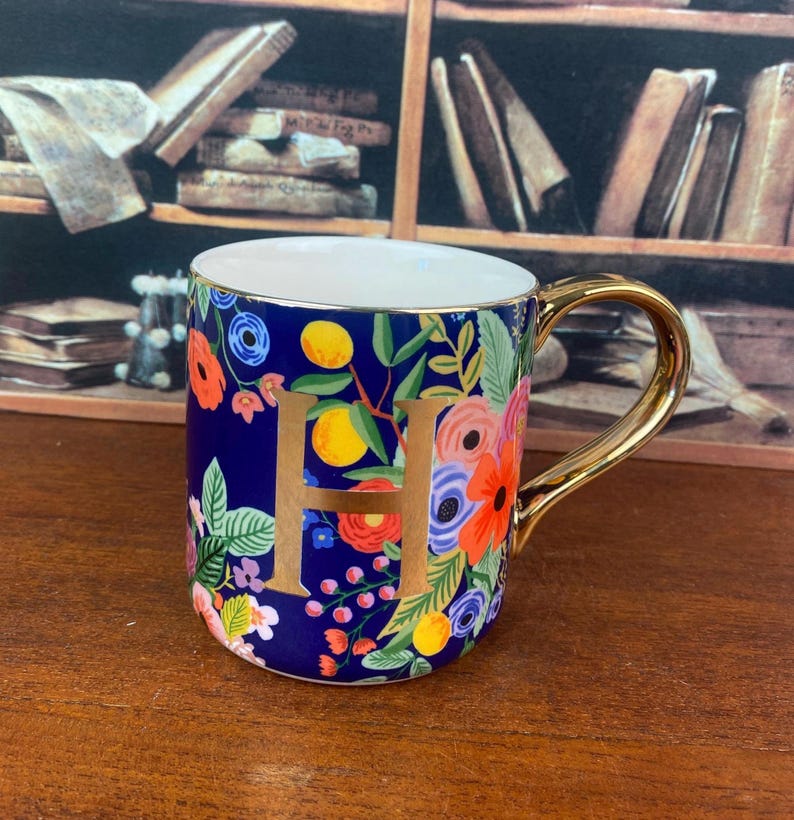 May include: A ceramic mug with a gold handle and the letter "H" in gold. The mug is decorated with a floral pattern in shades of blue, orange, pink, and green. The mug is sitting on a wooden surface.