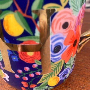May include: A ceramic mug with a floral design. The mug features a dark blue background with colorful flowers, including red, pink, and blue blooms, and yellow fruit. The handle and the letter "F" are gold.