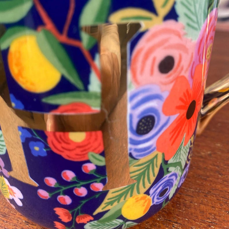 May include: A ceramic mug with a floral design. The mug features a dark blue background with colorful flowers, including red, pink, and blue blooms, and yellow fruit. The handle and the letter "F" are gold.