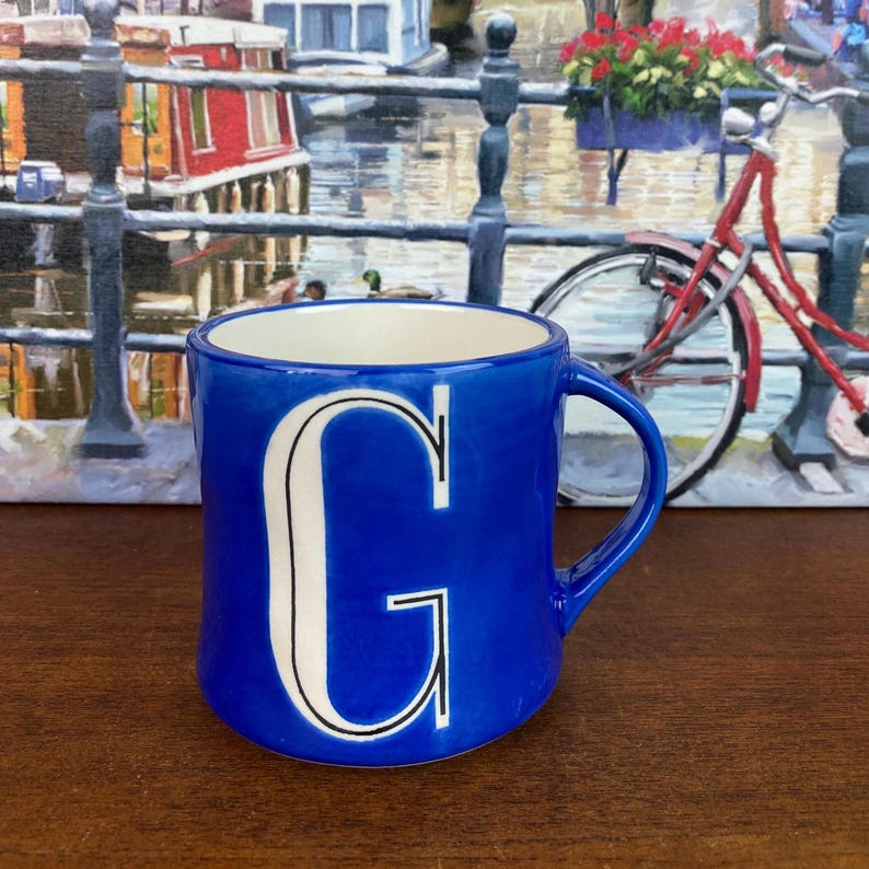 May include: A cobalt blue ceramic mug with a white interior and a large white letter "G" on the front. The mug has a curved handle and is set against a backdrop of a painting of a canal scene with a red bicycle.