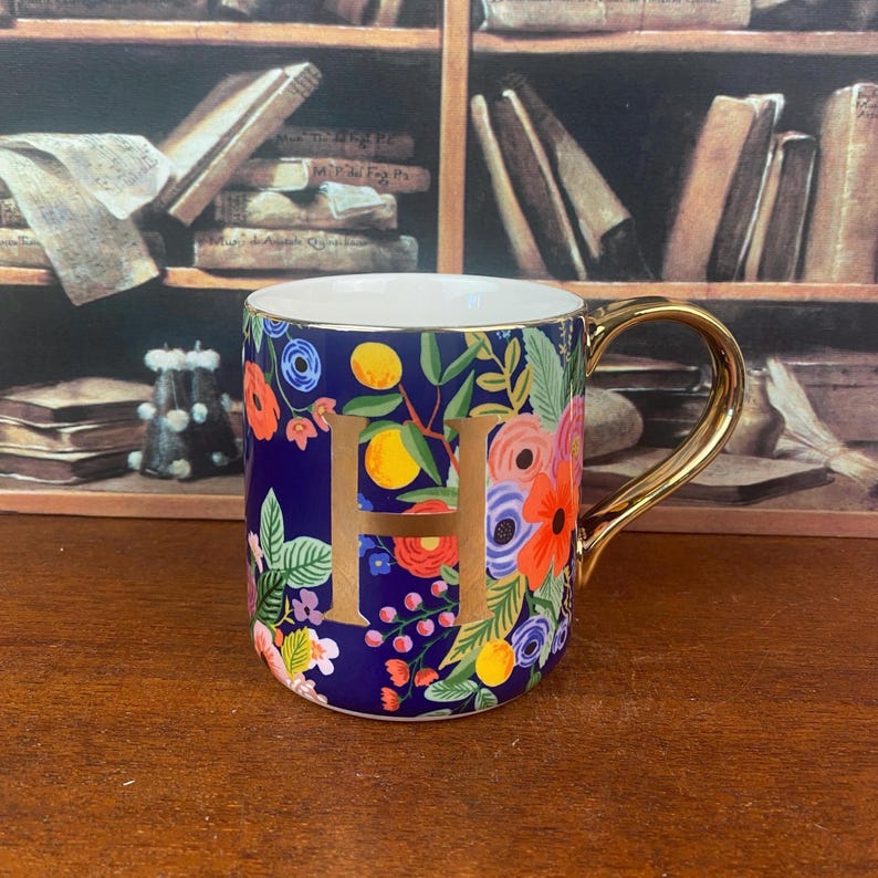 May include: A ceramic mug with a gold handle and the letter "H" in gold. The mug is decorated with colorful floral patterns on a dark blue background. The interior of the mug is white.