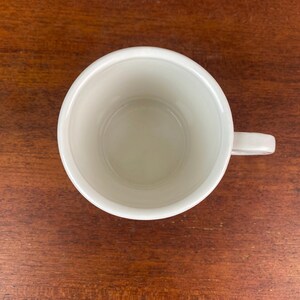 May include: Overhead view of a white ceramic mug with a handle. The mug is empty and sits on a dark brown wooden surface. The mug's interior is a light cream color.