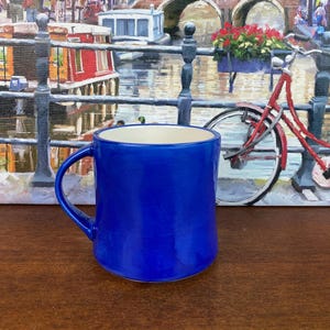 May include: A vibrant cobalt blue ceramic mug with a curved handle. The mug has a glossy finish and a cream interior. The background features a painting of a canal scene with a red bicycle.