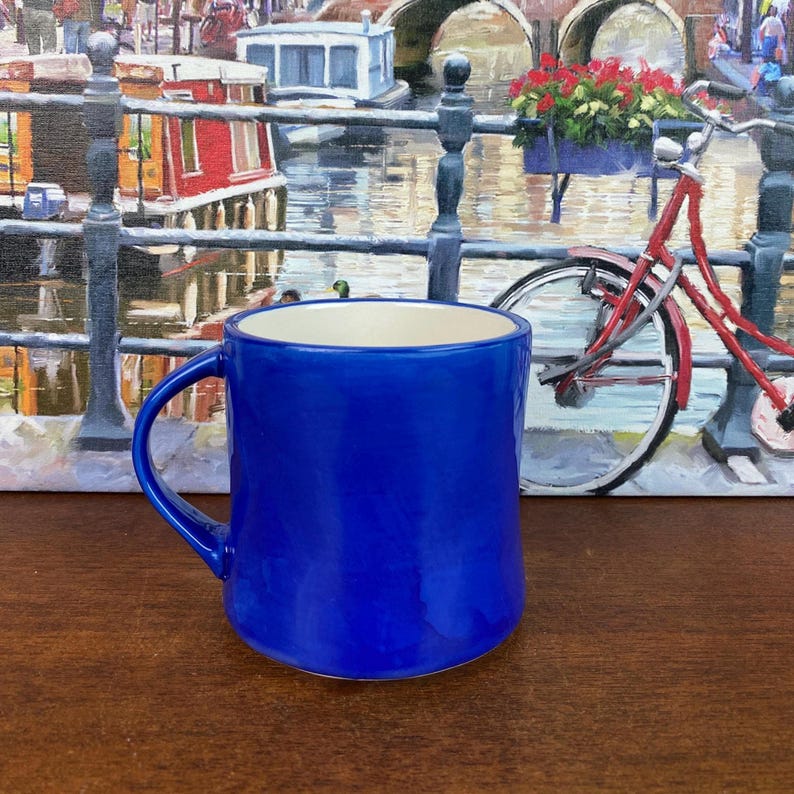 May include: A vibrant cobalt blue ceramic mug with a curved handle. The mug has a glossy finish and a cream interior. The background features a painting of a canal scene with a red bicycle.