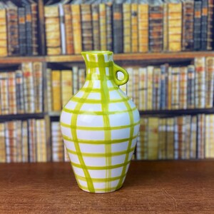 May include: A white ceramic vase with a green plaid pattern. The vase has a small handle and a narrow neck. The vase is on a wooden surface.