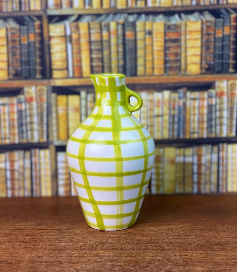 May include: A white ceramic vase with a green plaid pattern. The vase has a small handle and a narrow neck. The vase is on a wooden surface.