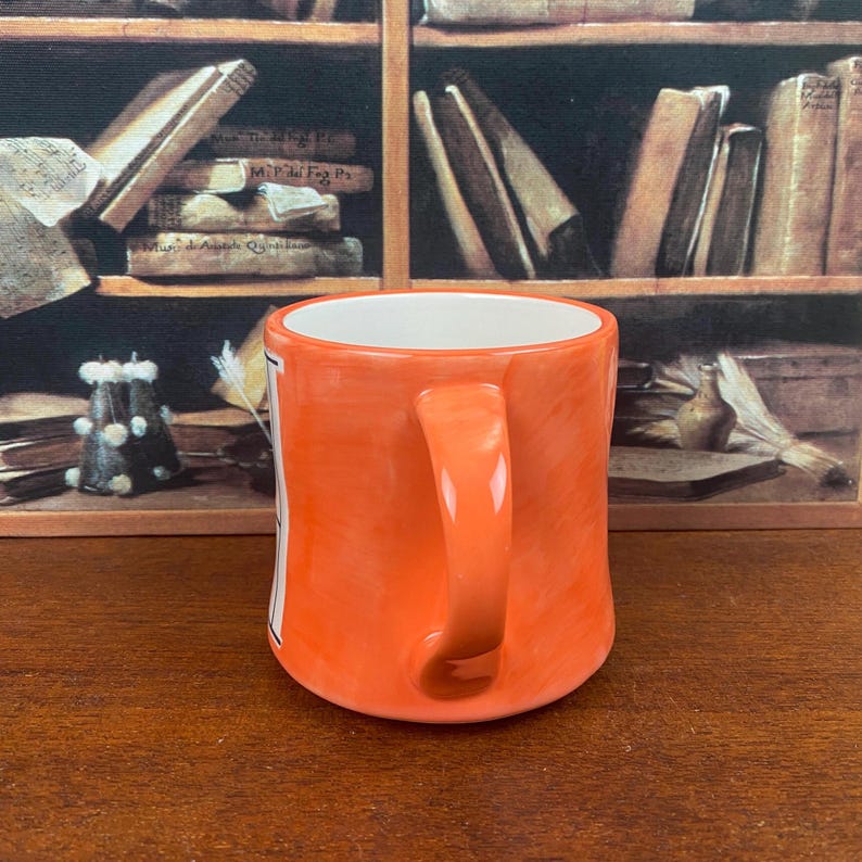 May include: An orange ceramic mug with a white interior and a curved handle. The mug has a glossy finish and a rectangular white label on the side. The background features a painting of a bookshelf.