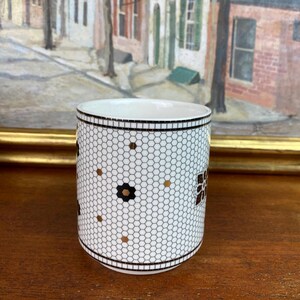 May include: White ceramic mug with a black and gold geometric pattern. The pattern resembles a honeycomb with small black flowers and gold dots.