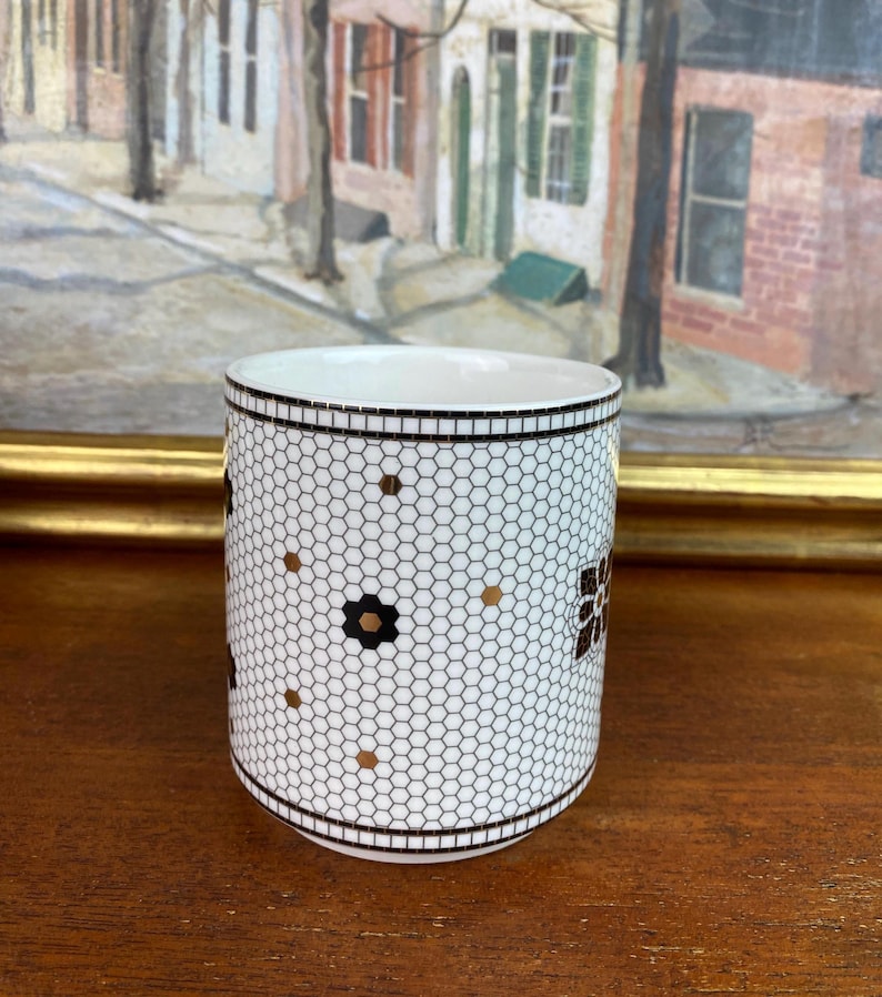 May include: White ceramic mug with a black and gold geometric pattern. The pattern resembles a honeycomb with small black flowers and gold dots.