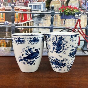 May include: Two white ceramic cups featuring blue hand-painted patterns. The left cup displays a bird perched on a branch, while the right cup is decorated with an all-over floral motif. Both cups have a tapered shape and a blue rim detail.