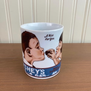 May include: White ceramic mug with a vintage illustration of two people about to kiss, holding a Hershey's chocolate. The mug has the text "A Kiss for you" and "HERSHEY'S MILK CHOCOLATE".