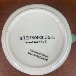 May include: Bottom view of a white ceramic mug with a light green handle. The text "ANTHROPOLOGIE hand painted" is printed in black. Additional text indicates the mug is dishwasher and microwave safe, made in China.