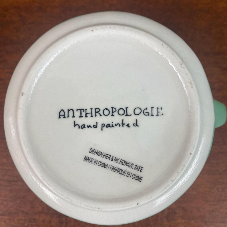 May include: Bottom view of a white ceramic mug with a light green handle. The text "ANTHROPOLOGIE hand painted" is printed in black. Additional text indicates the mug is dishwasher and microwave safe, made in China.